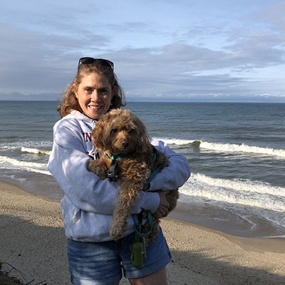 Photo of Academic Advisor and Manager of the Neurobiology major Cathy Auger on the beach holding a light brown small dog.